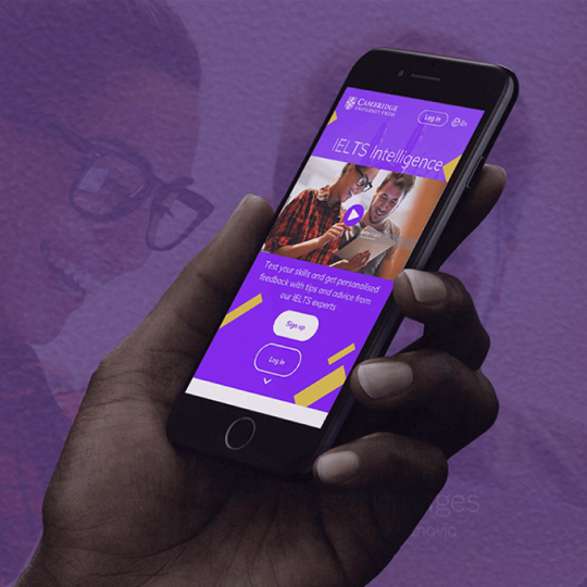 Photograph of a hand holding a smartphone displaying a purple IELTS learning app interface